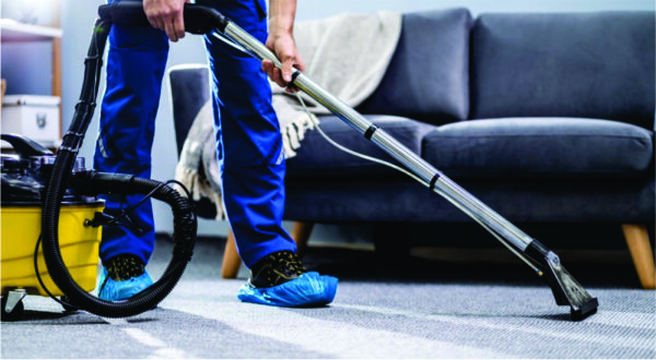 A person wearing blue pants and shoe covers uses a vacuum cleaner on a carpet in a living room with a gray sofa and a light-colored blanket.