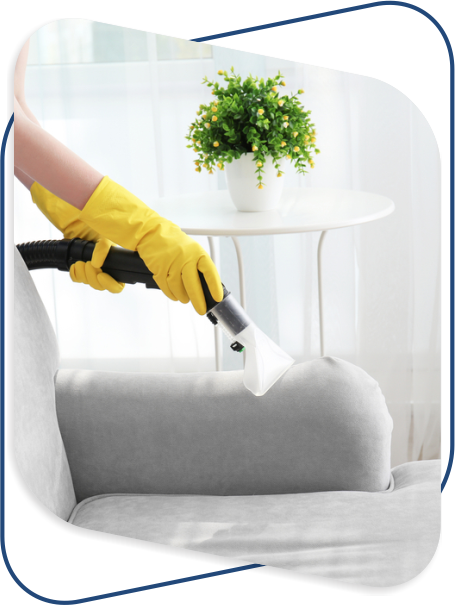 Person in yellow gloves uses a vacuum cleaner on a gray sofa near a table with a potted plant, conveying a sense of cleanliness and freshness.