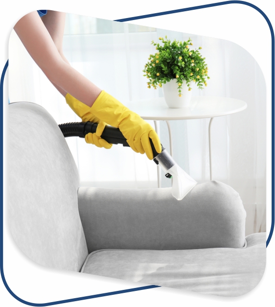 A person wearing yellow gloves is using a vacuum to clean a gray sofa in a bright room. A plant on a round table adds a fresh touch.
