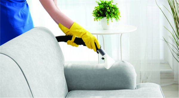 A person in yellow gloves uses a vacuum cleaner on a light grey sofa in a bright, tidy room. Nearby, a small plant sits on a white table.