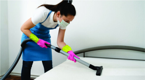 A woman in a blue apron and mask is vacuuming a mattress. She wears green and pink gloves, focusing intently. The room has a clean, minimalist look.