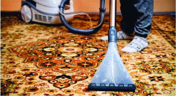 Person cleaning an ornate carpet with a carpet cleaner. The vacuum is visible, and the person is wearing patterned socks, suggesting a casual, homey atmosphere.