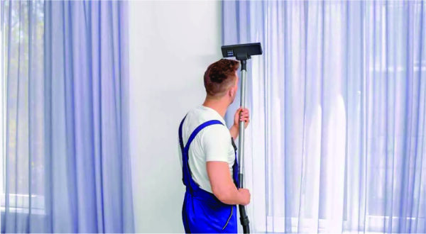A person in blue overalls stands facing a window, using a vacuum to clean the curtains. The setting is bright, conveying a sense of cleanliness and order.