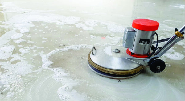 A floor cleaning machine with a red top scrubs a soapy, wet surface. White foam spreads across the shiny floor, suggesting active cleaning.