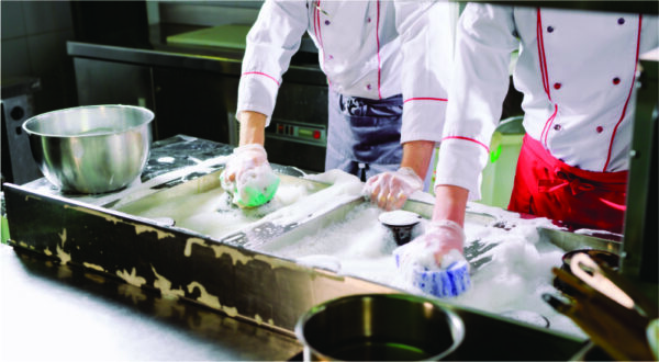 Chefs in white uniforms scrub a large kitchen sink filled with soapy water, using sponges. The scene conveys teamwork and cleanliness.