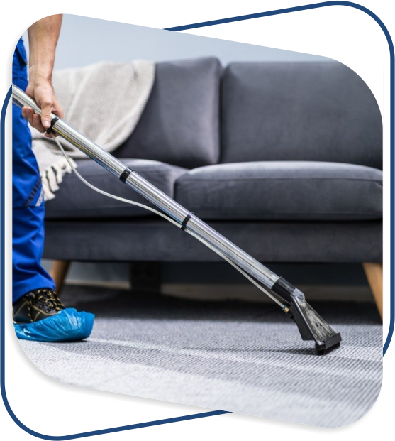 A person in blue pants and shoe covers uses a carpet cleaning machine on a gray carpet. A dark gray sofa with a folded blanket is in the background.
